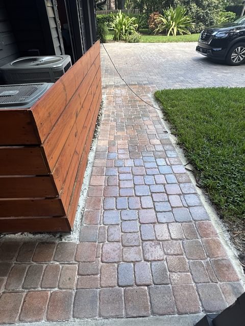 Camino de adoquines rojizos y grises junto a una celosía horizontal de cedro que rodea dos equipos de aire acondicionado, con césped a un lado y una entrada de adoquines al fondo, en Tampa Bay