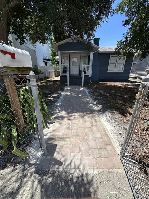 Camino corto de adoquines rojo ladrillo que va desde un portón de malla hasta los escalones de una casa azul, con tierra rastrillada a ambos lados, en Tampa Bay
