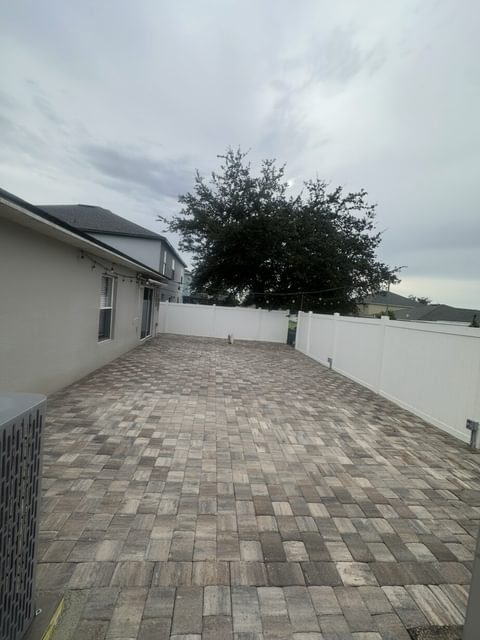 Gran patio de adoquines de varios tonos a lo largo de una casa hasta una cerca de vinilo blanca, con un árbol grande al fondo, en Tampa Bay
